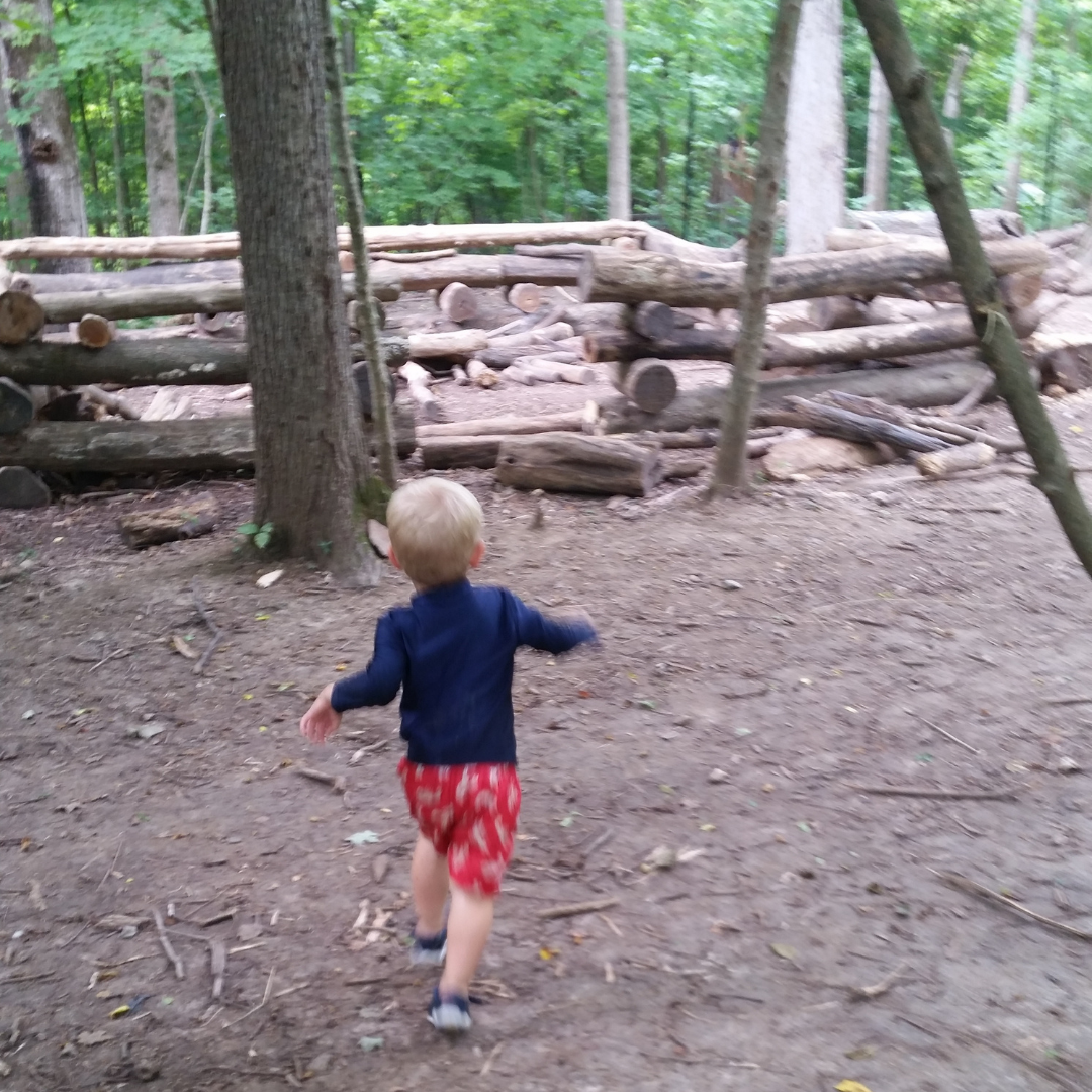 A young blonde boy in a long sleeved navy top and red shorts runs toward a log fort inside the wooded area of the Nature PlayScape. 